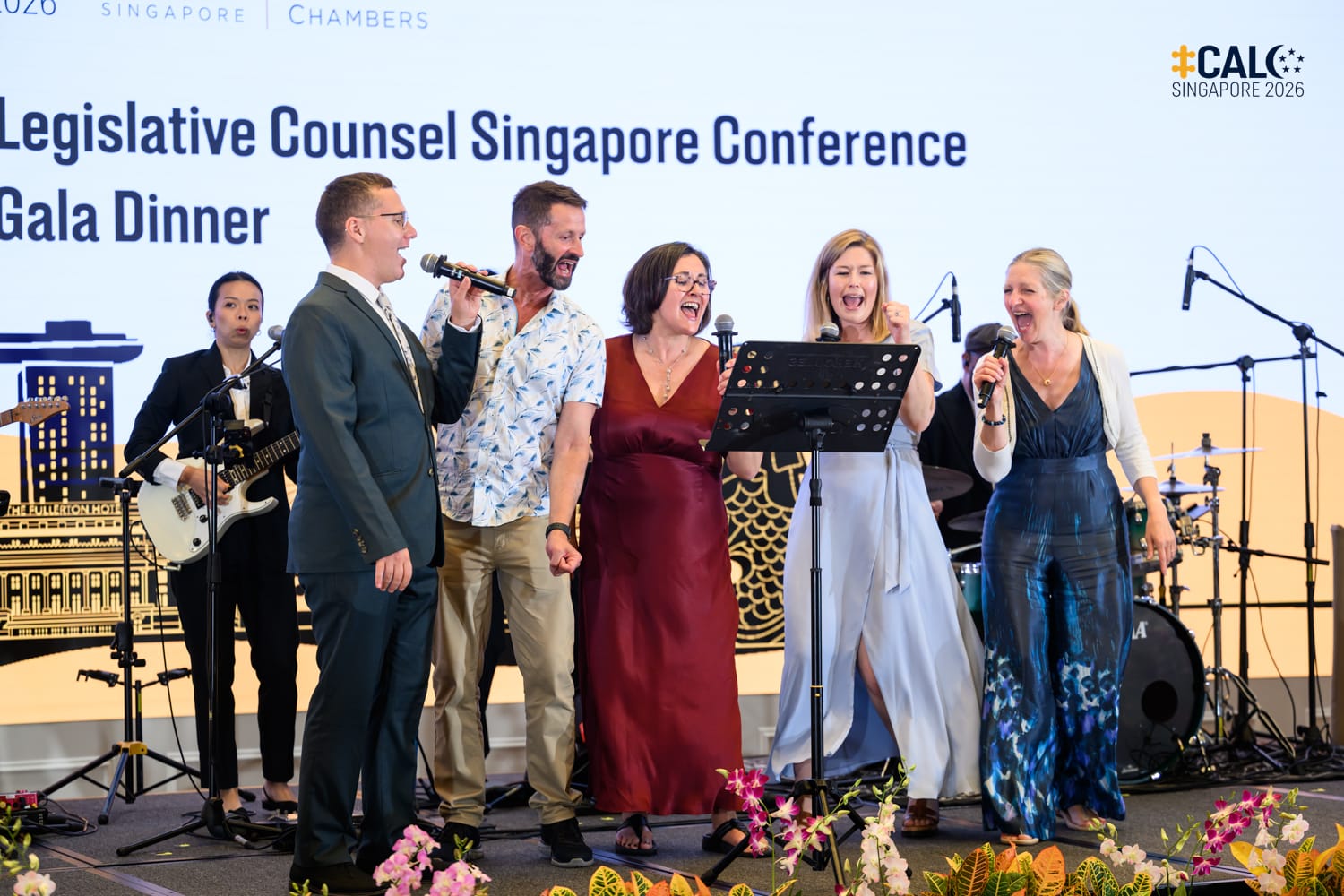 Five delegates sing on stage together at the Conference Gala Dinner, accompanied by a guitarist from band 53A in the background.